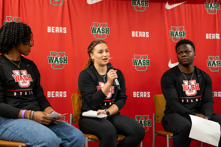 three students sit on chairs while one student speaks on microphone